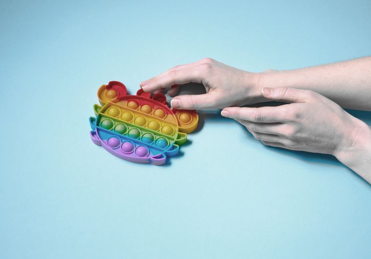 Hand protruding into the picture from the right, touching a crab-shaped Pop-it toy in rainbow colors lying on the table